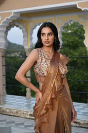 A woman poses on a rooftop terrace wearing a metallic bronze-toned draped saree featuring a voluminous ruffled border. The ensemble includes a sleeveless, sheer-style blouse heavily embellished with gold and silver floral embroidery. She stands before a backdrop of traditional white scalloped arches and lush green trees in the distance.