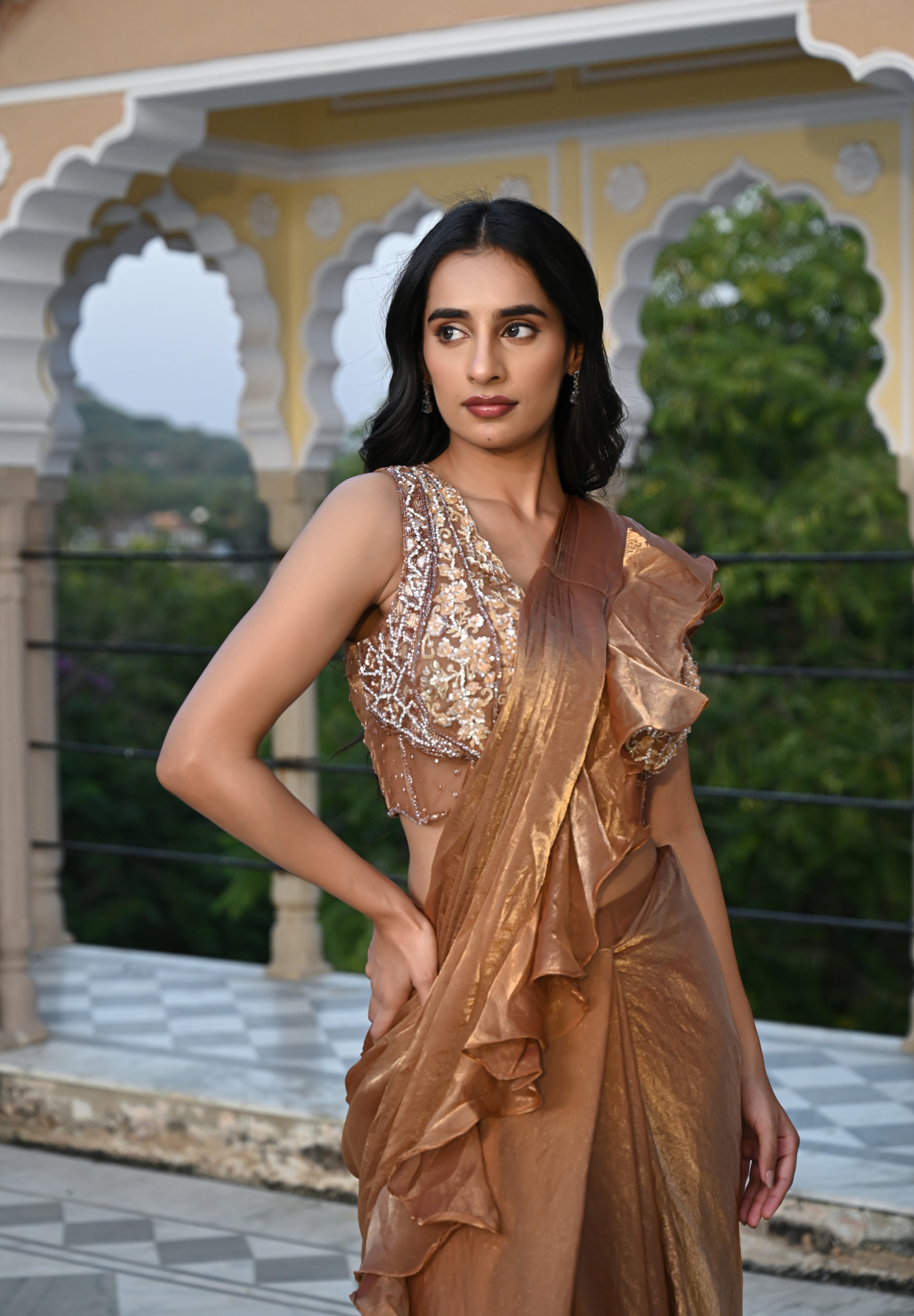 A woman poses on a rooftop terrace wearing a metallic bronze-toned draped saree featuring a voluminous ruffled border. The ensemble includes a sleeveless, sheer-style blouse heavily embellished with gold and silver floral embroidery. She stands before a backdrop of traditional white scalloped arches and lush green trees in the distance.