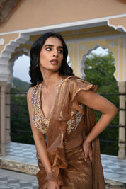 A woman poses on a rooftop terrace wearing a metallic bronze-toned draped saree featuring a voluminous ruffled border. The ensemble includes a sleeveless, sheer-style blouse heavily embellished with gold and silver floral embroidery. She stands before a backdrop of traditional white scalloped arches and lush green trees in the distance.