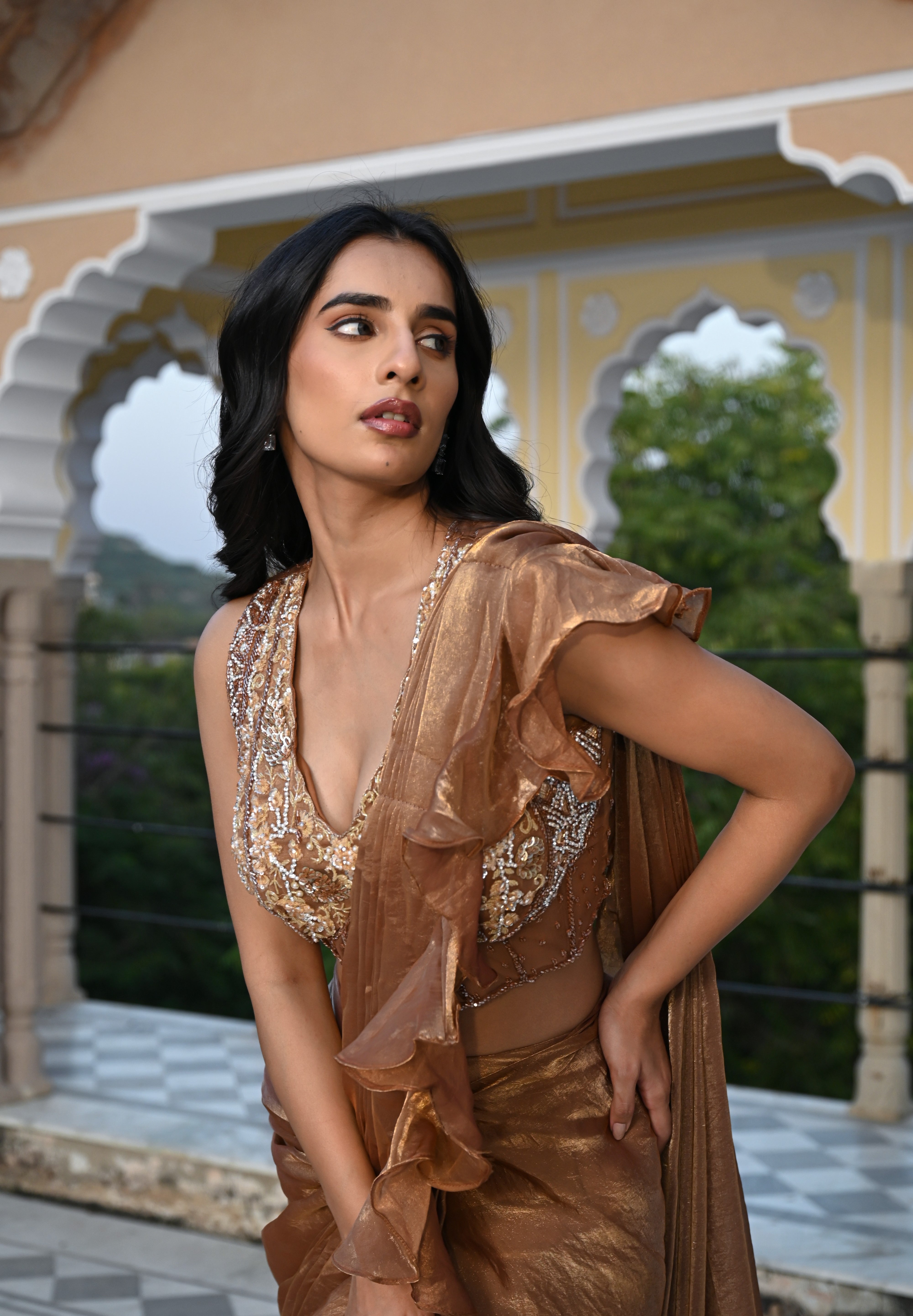A woman poses on a rooftop terrace wearing a metallic bronze-toned draped saree featuring a voluminous ruffled border. The ensemble includes a sleeveless, sheer-style blouse heavily embellished with gold and silver floral embroidery. She stands before a backdrop of traditional white scalloped arches and lush green trees in the distance.