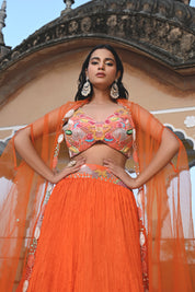 A woman stands on a rooftop wearing a vibrant orange crushed-silk lehenga. The look features a multicolored embroidered crop top with pink, yellow, and green floral details, paired with a sheer orange cape that has delicate silver embroidery along the edges. The background features the ornate domes of a traditional Indian building against a clear sky.