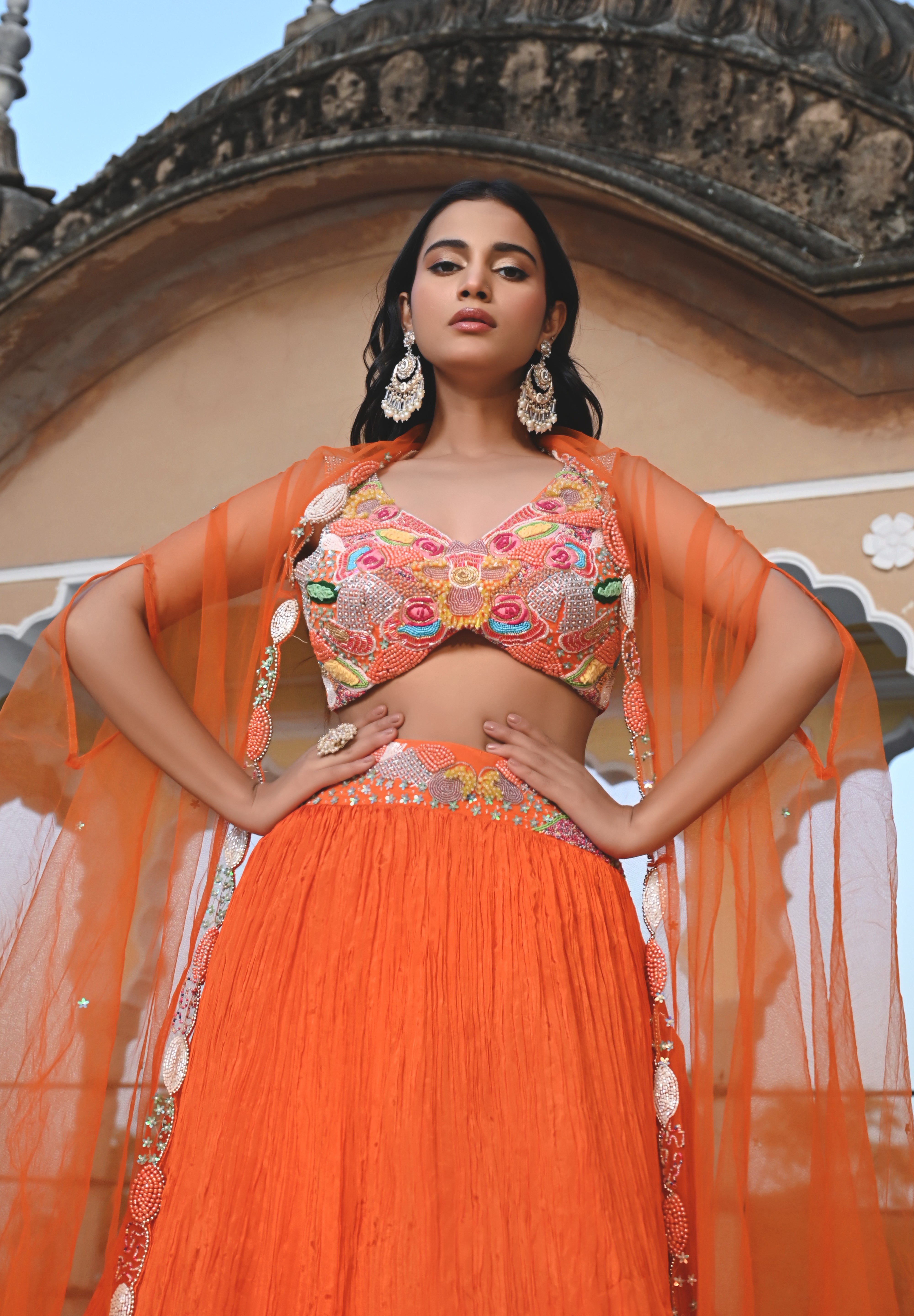 A woman stands on a rooftop wearing a vibrant orange crushed-silk lehenga. The look features a multicolored embroidered crop top with pink, yellow, and green floral details, paired with a sheer orange cape that has delicate silver embroidery along the edges. The background features the ornate domes of a traditional Indian building against a clear sky.