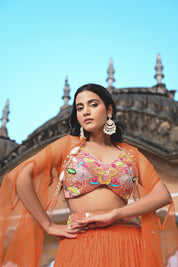 A woman stands on a rooftop wearing a vibrant orange crushed-silk lehenga. The look features a multicolored embroidered crop top with pink, yellow, and green floral details, paired with a sheer orange cape that has delicate silver embroidery along the edges. The background features the ornate domes of a traditional Indian building against a clear sky.