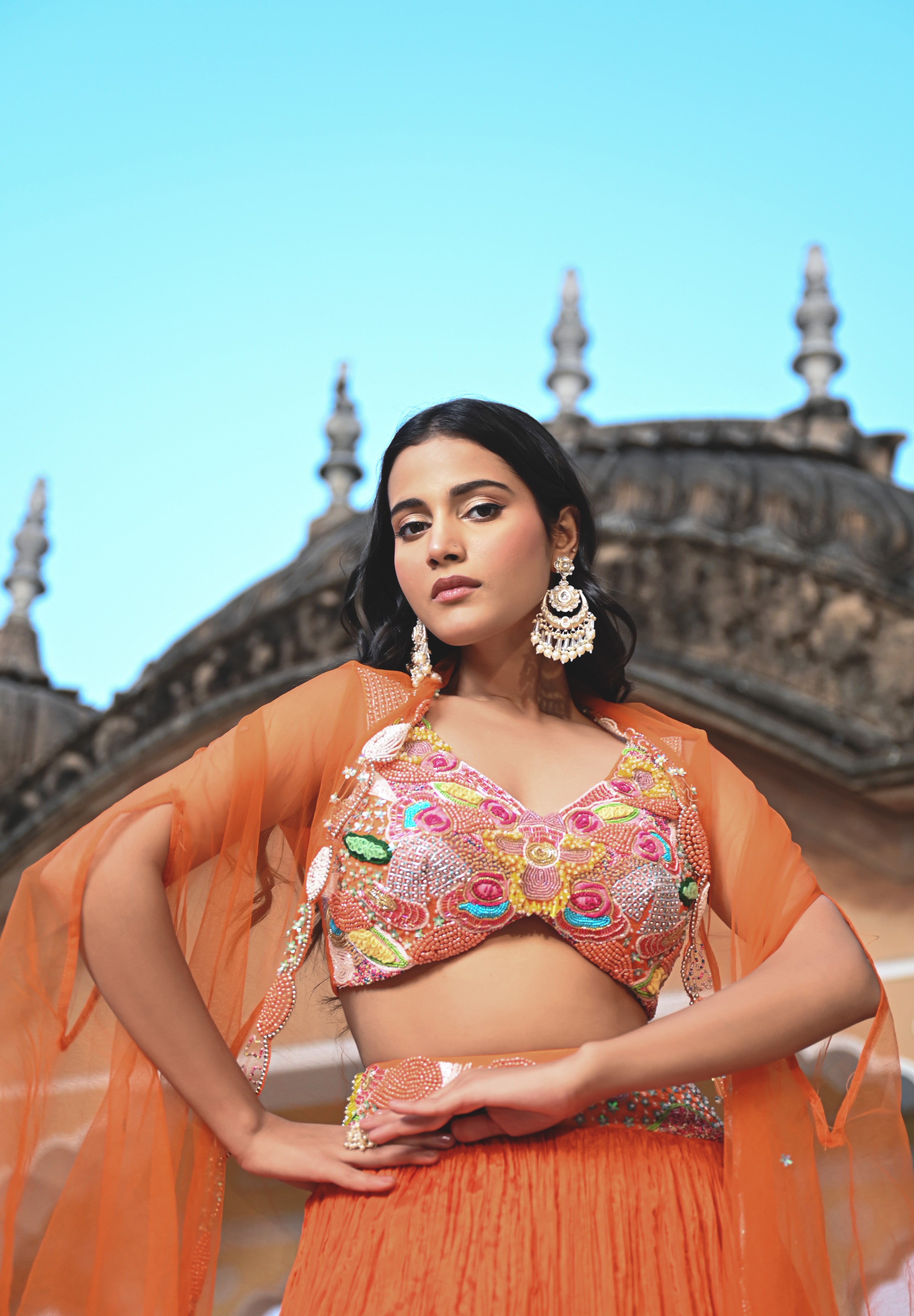 A woman stands on a rooftop wearing a vibrant orange crushed-silk lehenga. The look features a multicolored embroidered crop top with pink, yellow, and green floral details, paired with a sheer orange cape that has delicate silver embroidery along the edges. The background features the ornate domes of a traditional Indian building against a clear sky.