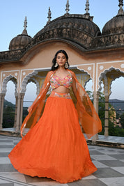 A woman stands on a rooftop wearing a vibrant orange crushed-silk lehenga. The look features a multicolored embroidered crop top with pink, yellow, and green floral details, paired with a sheer orange cape that has delicate silver embroidery along the edges. The background features the ornate domes of a traditional Indian building against a clear sky.