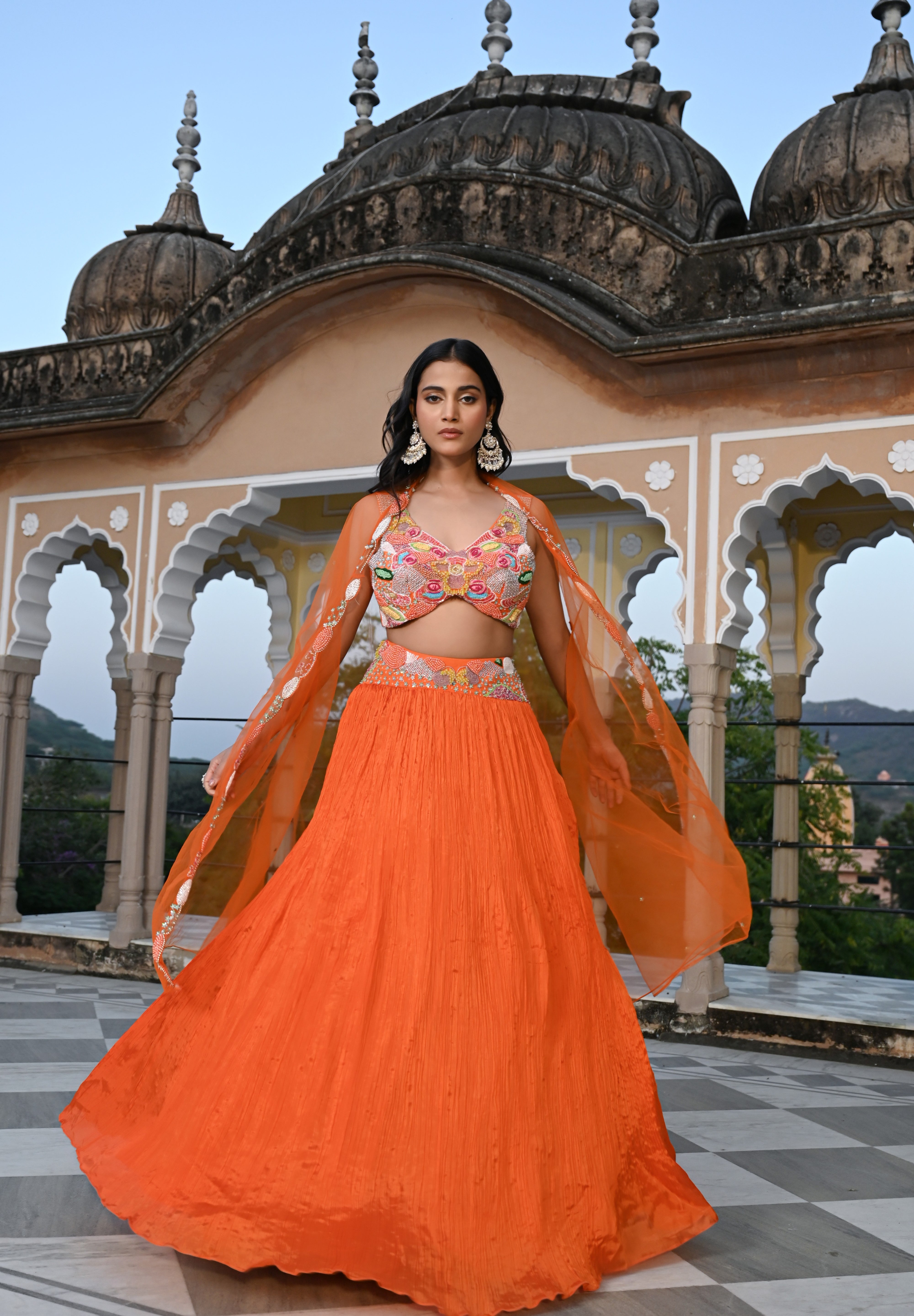A woman stands on a rooftop wearing a vibrant orange crushed-silk lehenga. The look features a multicolored embroidered crop top with pink, yellow, and green floral details, paired with a sheer orange cape that has delicate silver embroidery along the edges. The background features the ornate domes of a traditional Indian building against a clear sky.