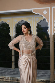 A woman poses at dusk under a traditional archway, wearing a champagne-gold draped skirt. The outfit features a matching embroidered bustier top with a long, shimmering pearl-bead fringe that covers the midriff, layered with a sheer three-quarter sleeve jacket. She is accessorized with large, ornate silver statement earrings.