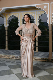 A woman poses at dusk under a traditional archway, wearing a champagne-gold draped skirt. The outfit features a matching embroidered bustier top with a long, shimmering pearl-bead fringe that covers the midriff, layered with a sheer three-quarter sleeve jacket. She is accessorized with large, ornate silver statement earrings.