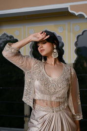 A woman poses at dusk under a traditional archway, wearing a champagne-gold draped skirt. The outfit features a matching embroidered bustier top with a long, shimmering pearl-bead fringe that covers the midriff, layered with a sheer three-quarter sleeve jacket. She is accessorized with large, ornate silver statement earrings.