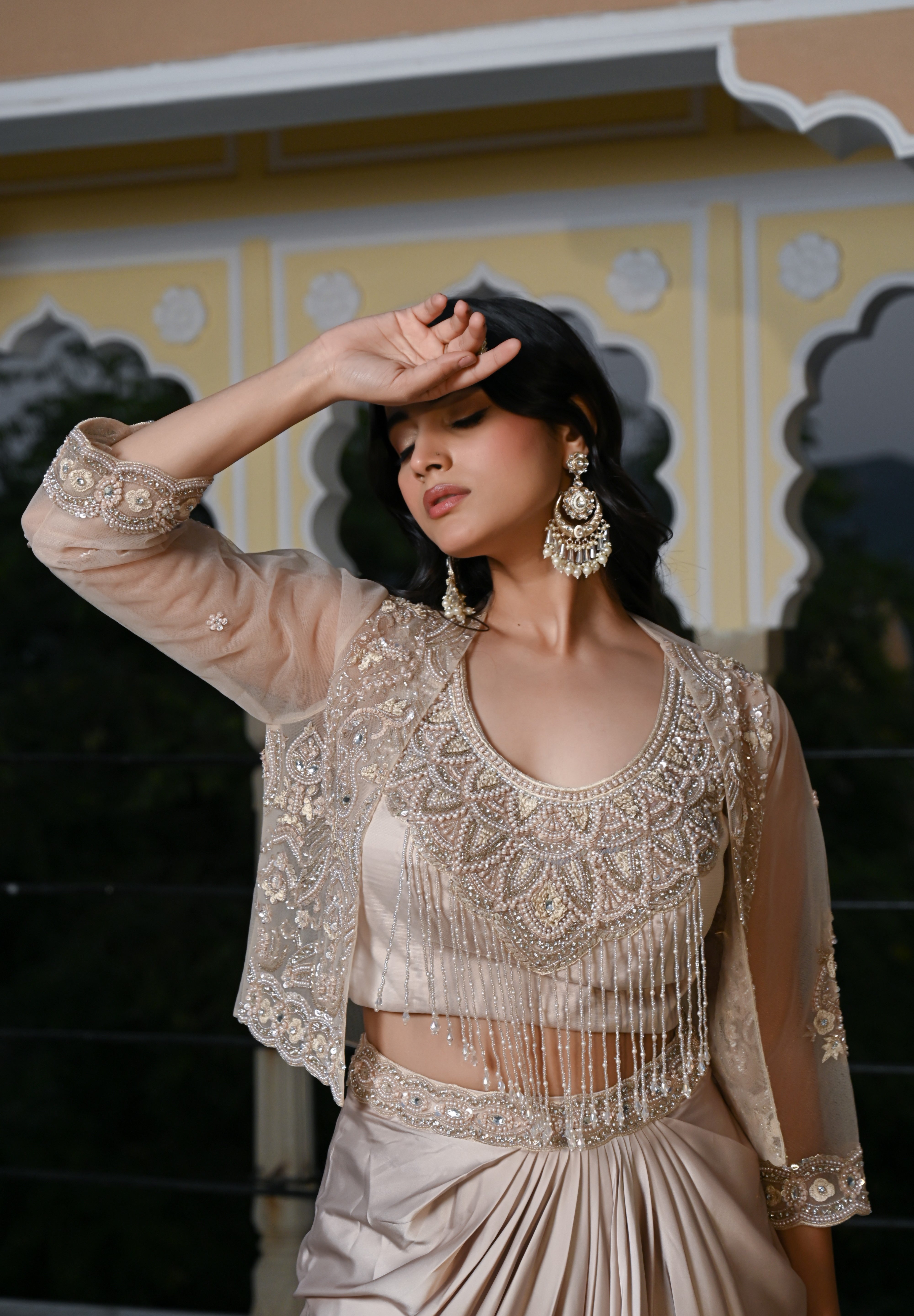 A woman poses at dusk under a traditional archway, wearing a champagne-gold draped skirt. The outfit features a matching embroidered bustier top with a long, shimmering pearl-bead fringe that covers the midriff, layered with a sheer three-quarter sleeve jacket. She is accessorized with large, ornate silver statement earrings.