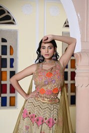 A woman poses near a stained glass archway wearing an olive-gold lehenga. The crop top features prominent orange and pink floral appliqués with a shell and bead fringe at the hem.