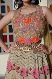 A woman poses near a stained glass archway wearing an olive-gold lehenga. The crop top features prominent orange and pink floral appliqués with a shell and bead fringe at the hem.