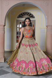 A woman poses near a stained glass archway wearing an olive-gold lehenga. The crop top features prominent orange and pink floral appliqués with a shell and bead fringe at the hem.