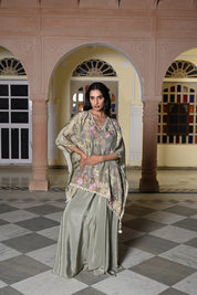 A woman stands in a traditional hall with arches wearing a sage green silk-style ensemble. It features a loose-fitting kaftan-style top with colorful floral embroidery and a white pom-pom trim, paired with a matching