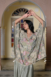 A woman stands in a traditional hall with arches wearing a sage green silk-style ensemble. It features a loose-fitting kaftan-style top with colorful floral embroidery and a white pom-pom trim, paired with a matching