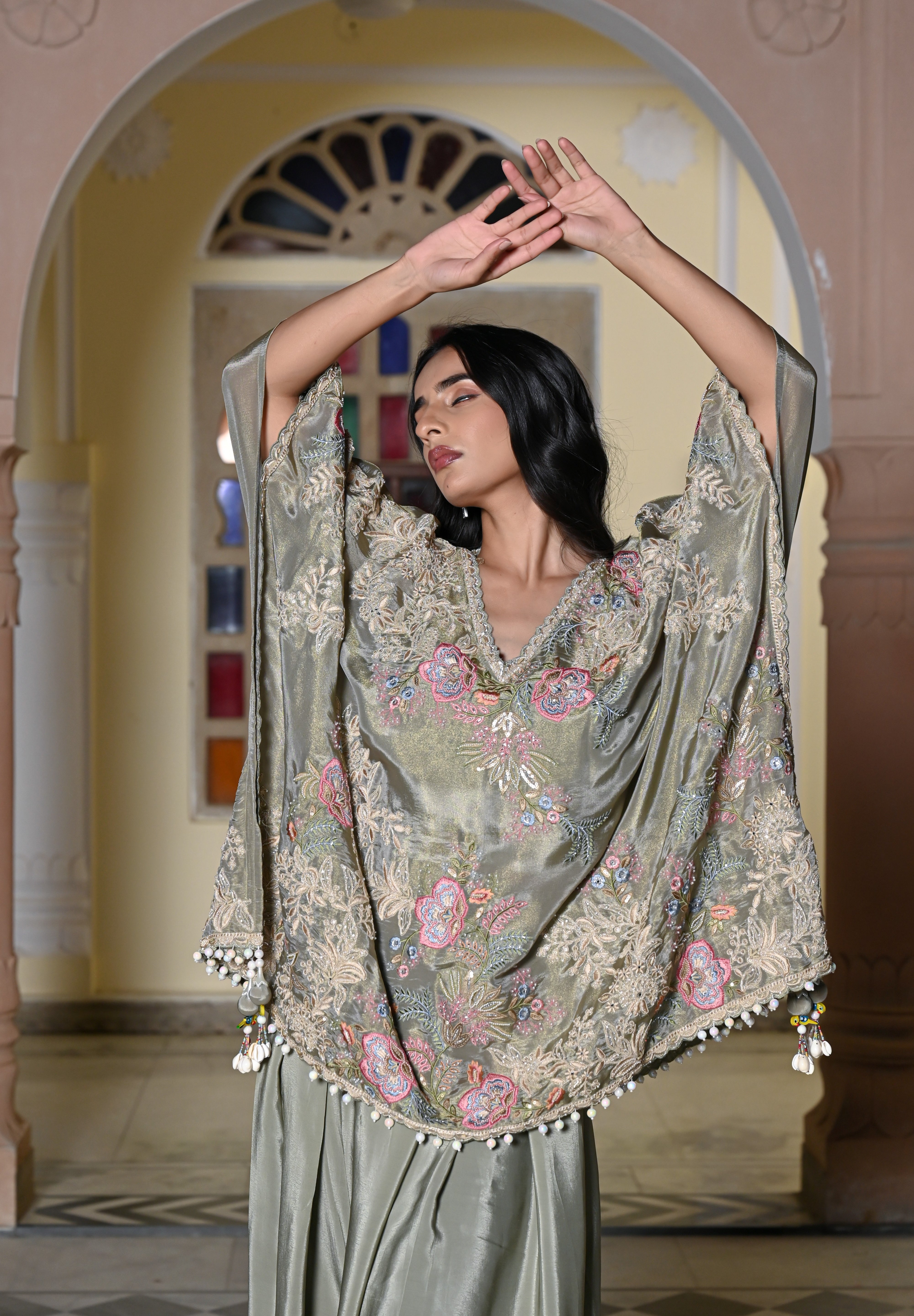 A woman stands in a traditional hall with arches wearing a sage green silk-style ensemble. It features a loose-fitting kaftan-style top with colorful floral embroidery and a white pom-pom trim, paired with a matching