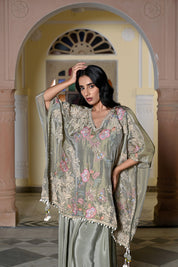 A woman stands in a traditional hall with arches wearing a sage green silk-style ensemble. It features a loose-fitting kaftan-style top with colorful floral embroidery and a white pom-pom trim, paired with a matching