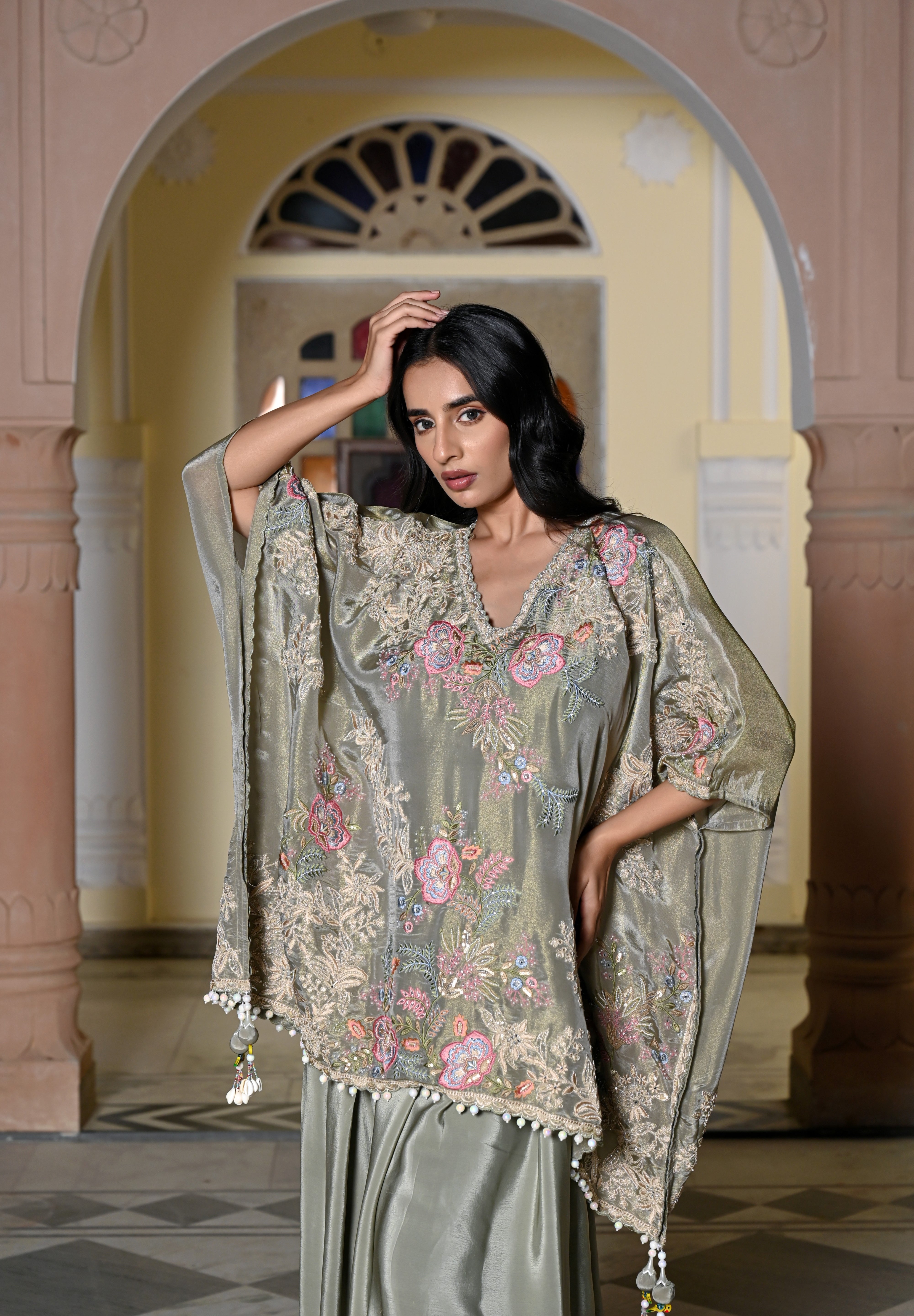 A woman stands in a traditional hall with arches wearing a sage green silk-style ensemble. It features a loose-fitting kaftan-style top with colorful floral embroidery and a white pom-pom trim, paired with a matching