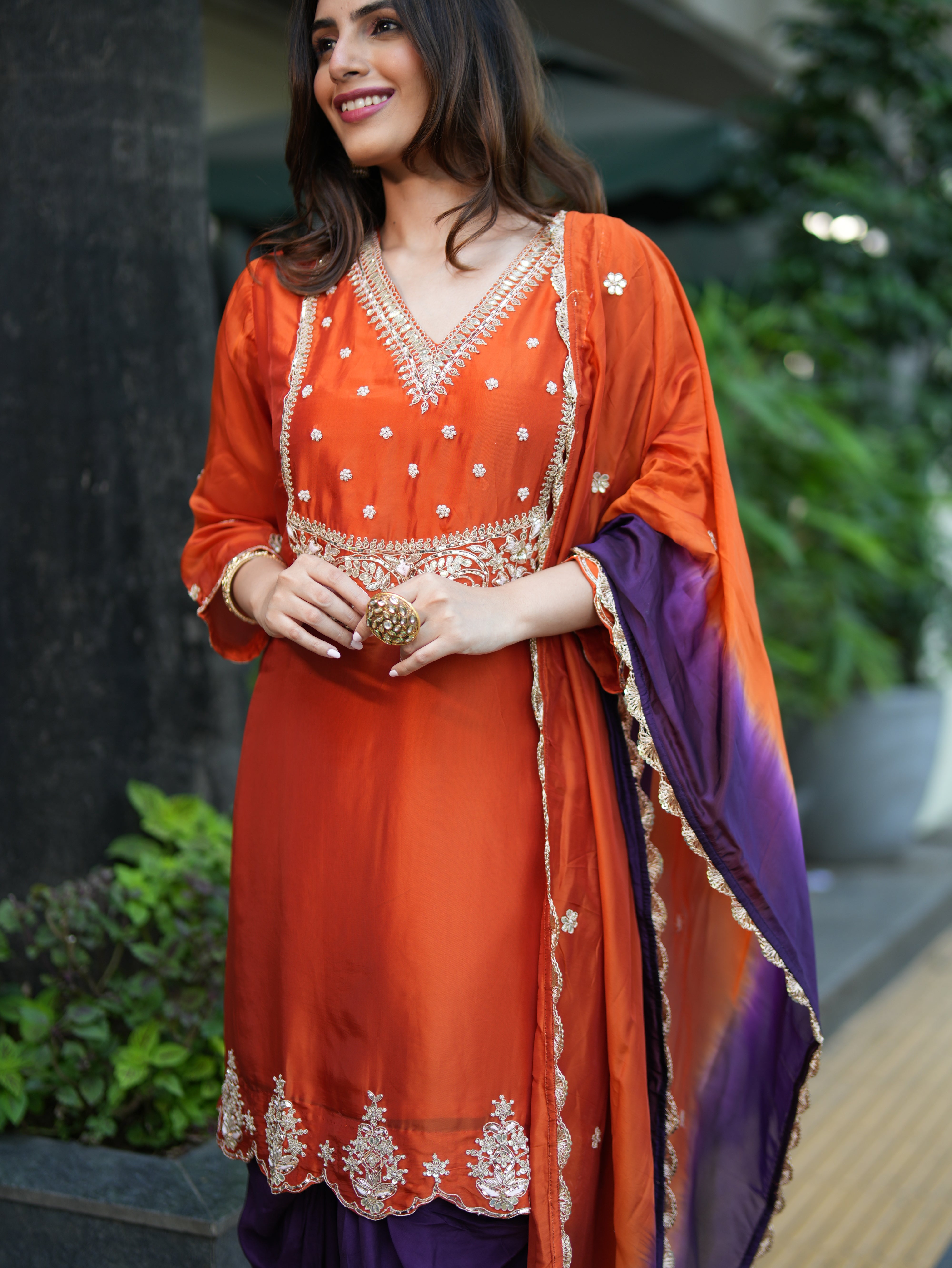 Woman wearing an orange and purple traditional outfit with embroidery, standing outdoors.