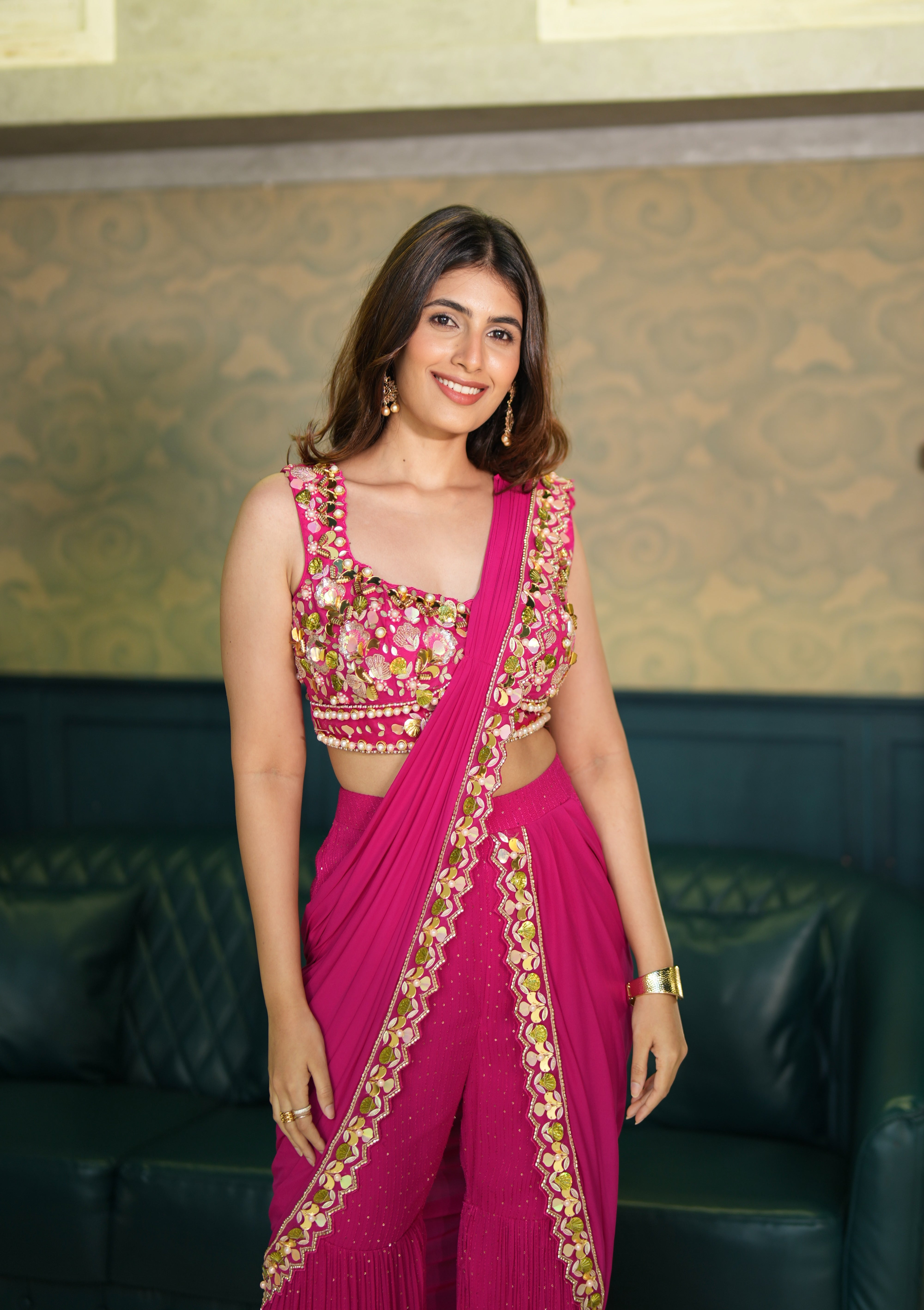 Woman in a pink indo western saree with gold embroidery standing against a patterned wall.