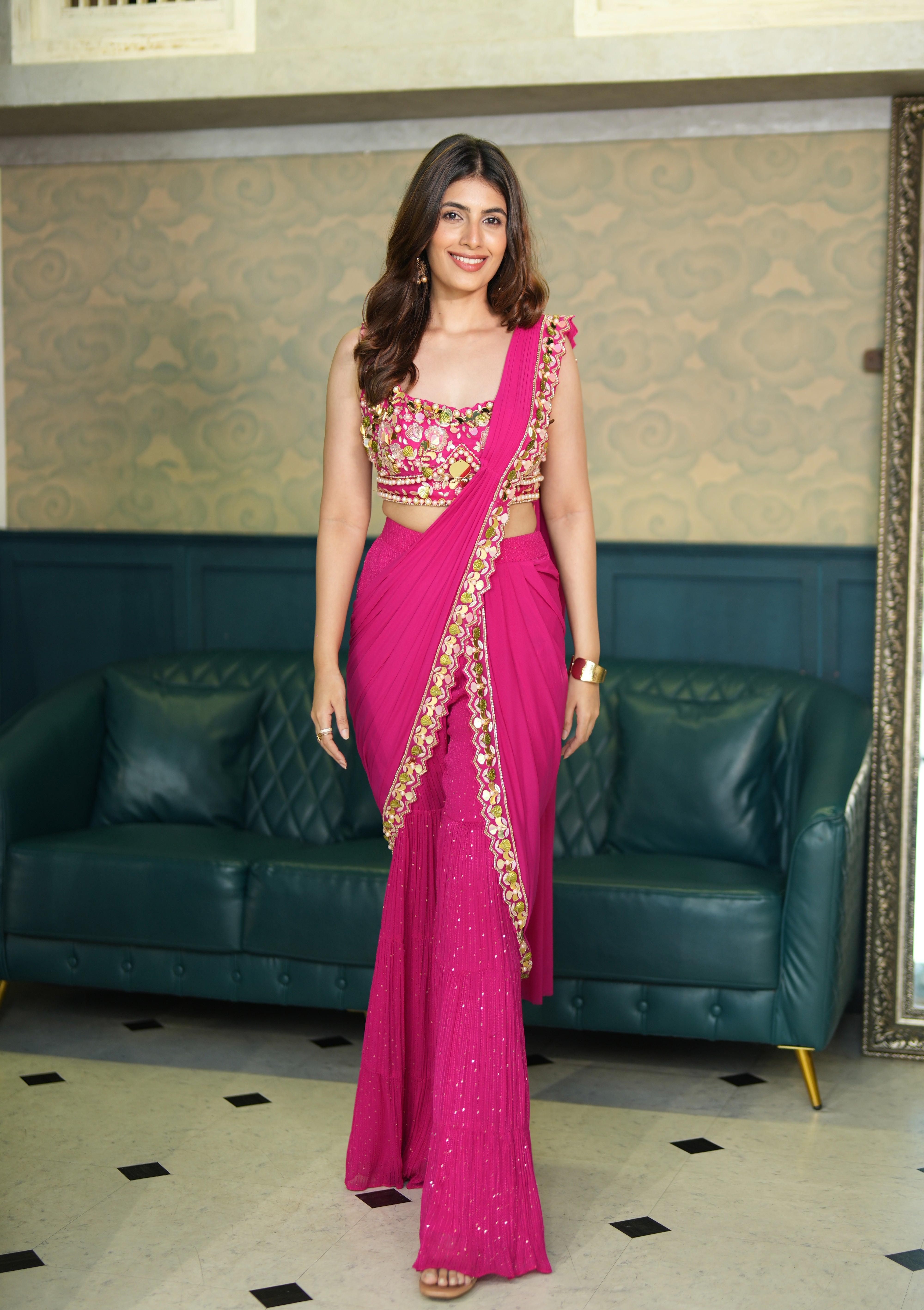Woman in a pink indo western saree standing in a room with green sofa and patterned wall.