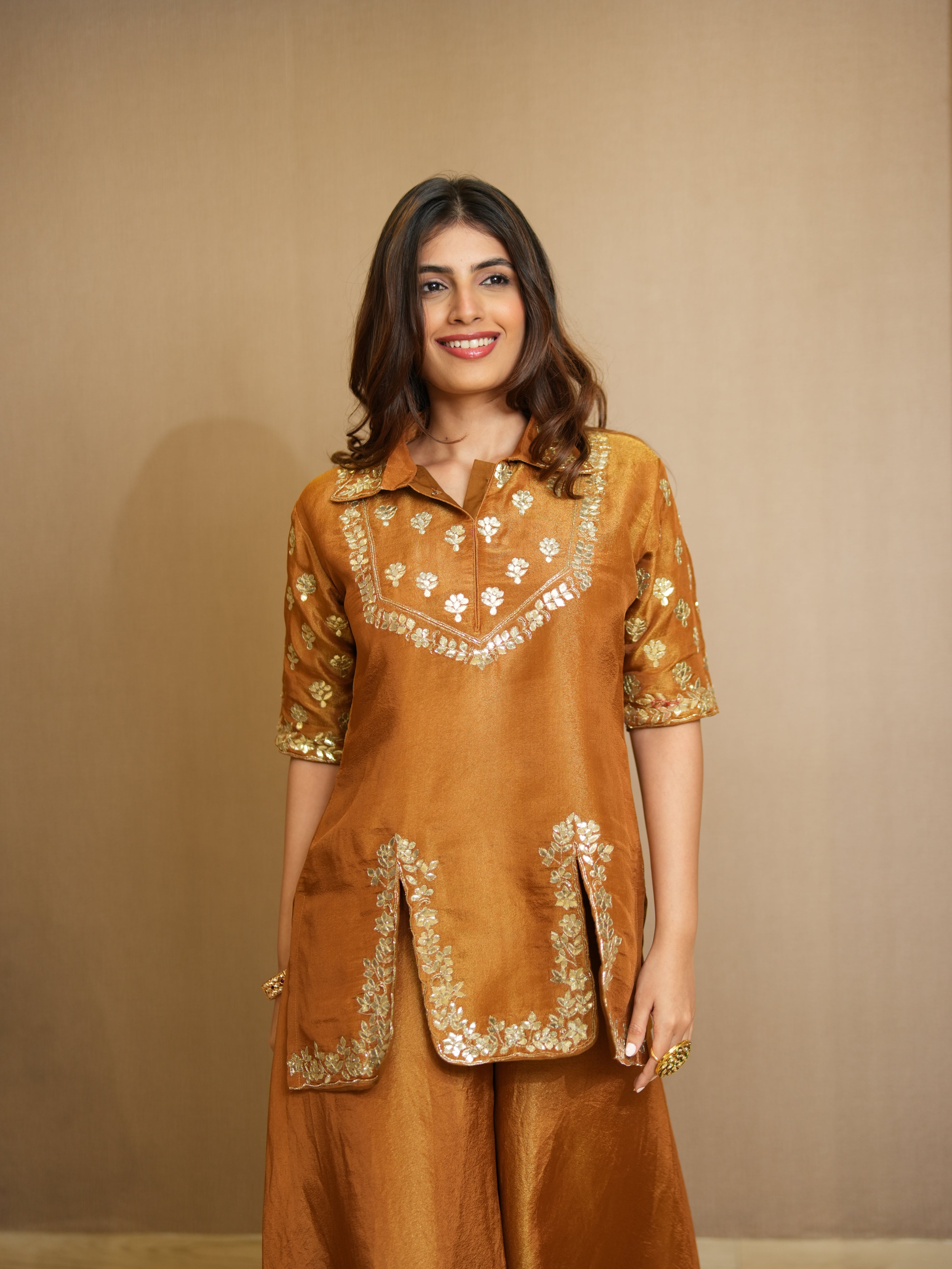 Woman wearing a mustard traditional outfit with gold embroidery against a beige background
