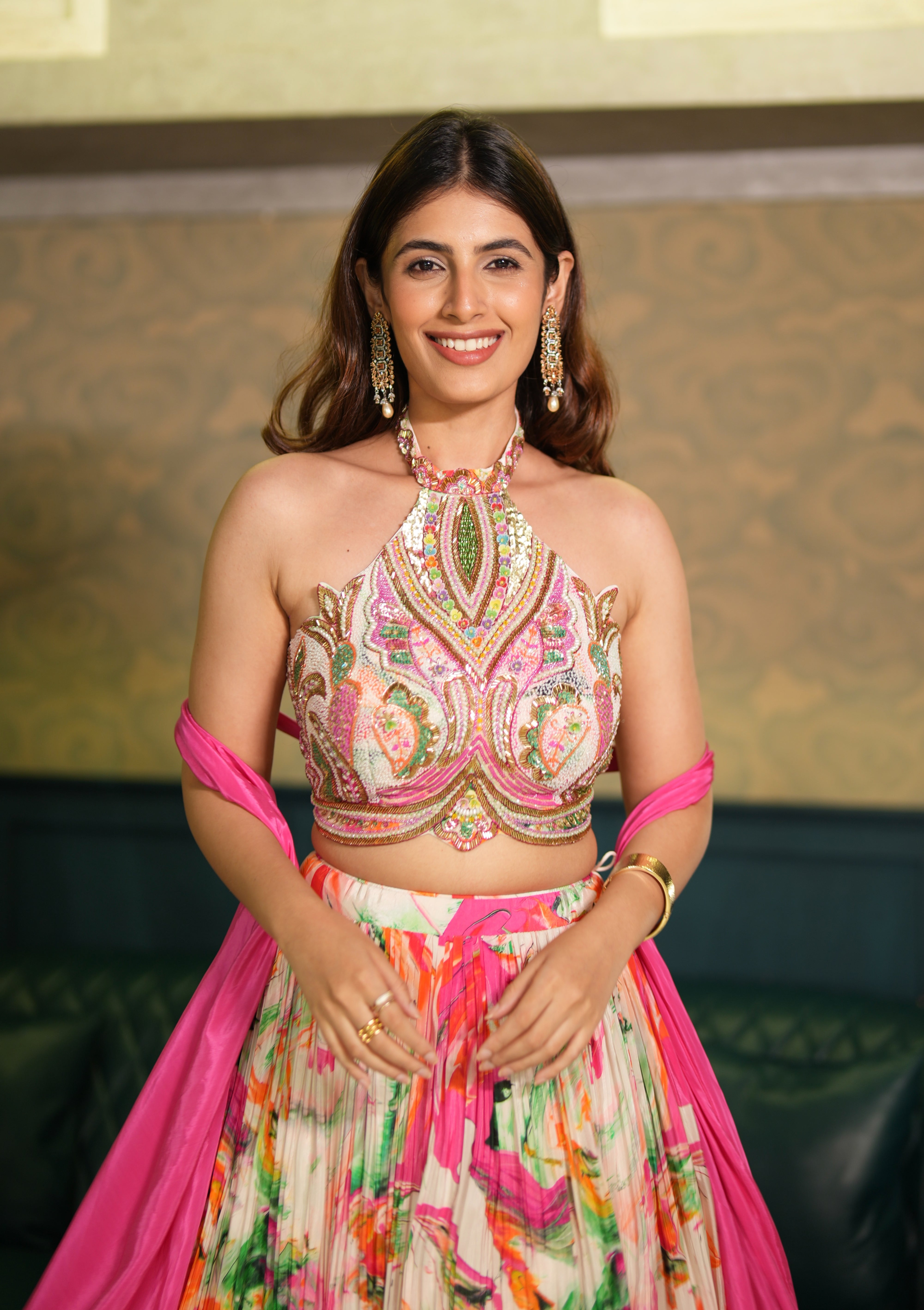 Woman in a colorful traditional outfit with pink dupatta against a patterned wall.