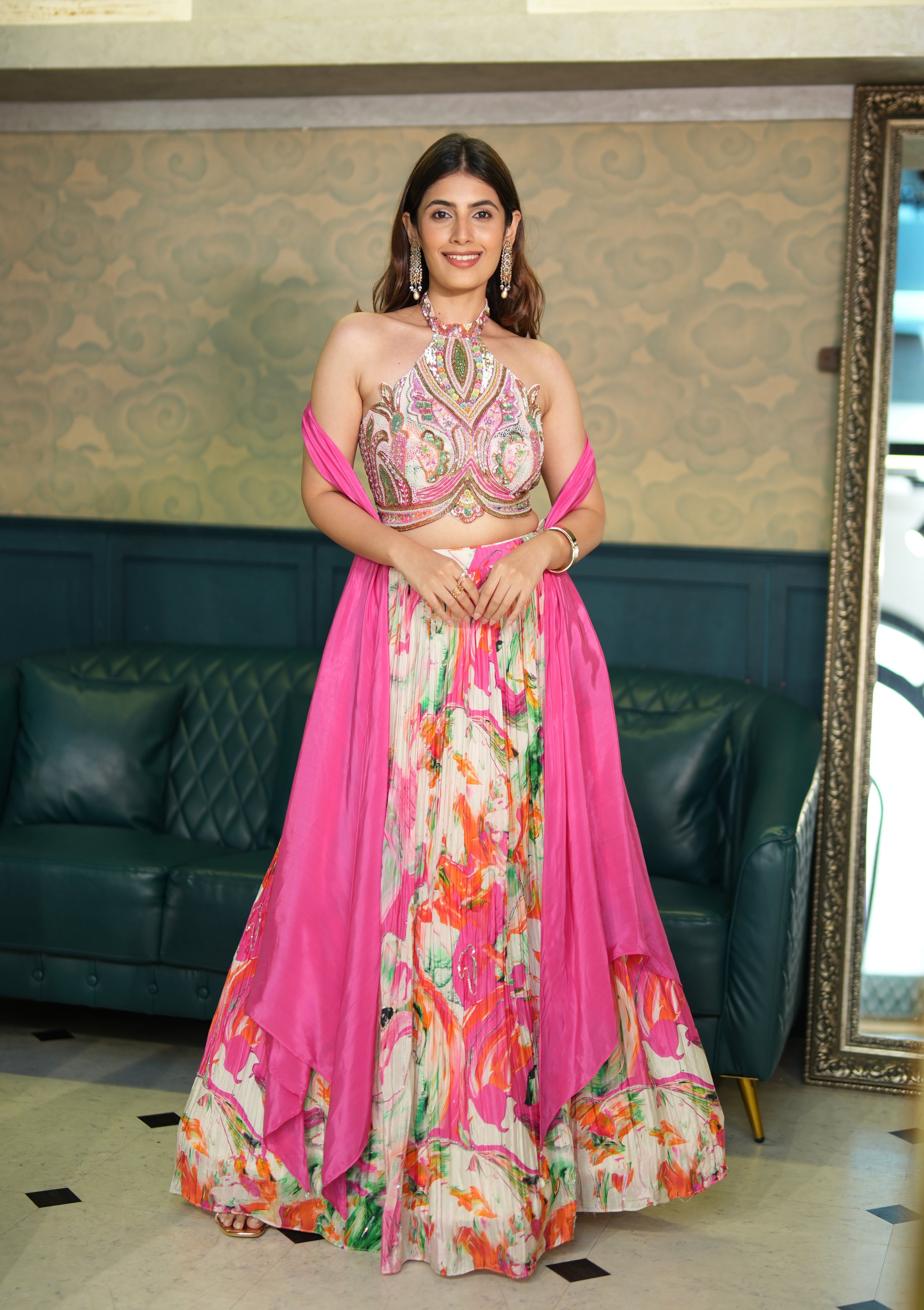 Woman in a floral and pink traditional outfit standing in a room with a green sofa and patterned wall.