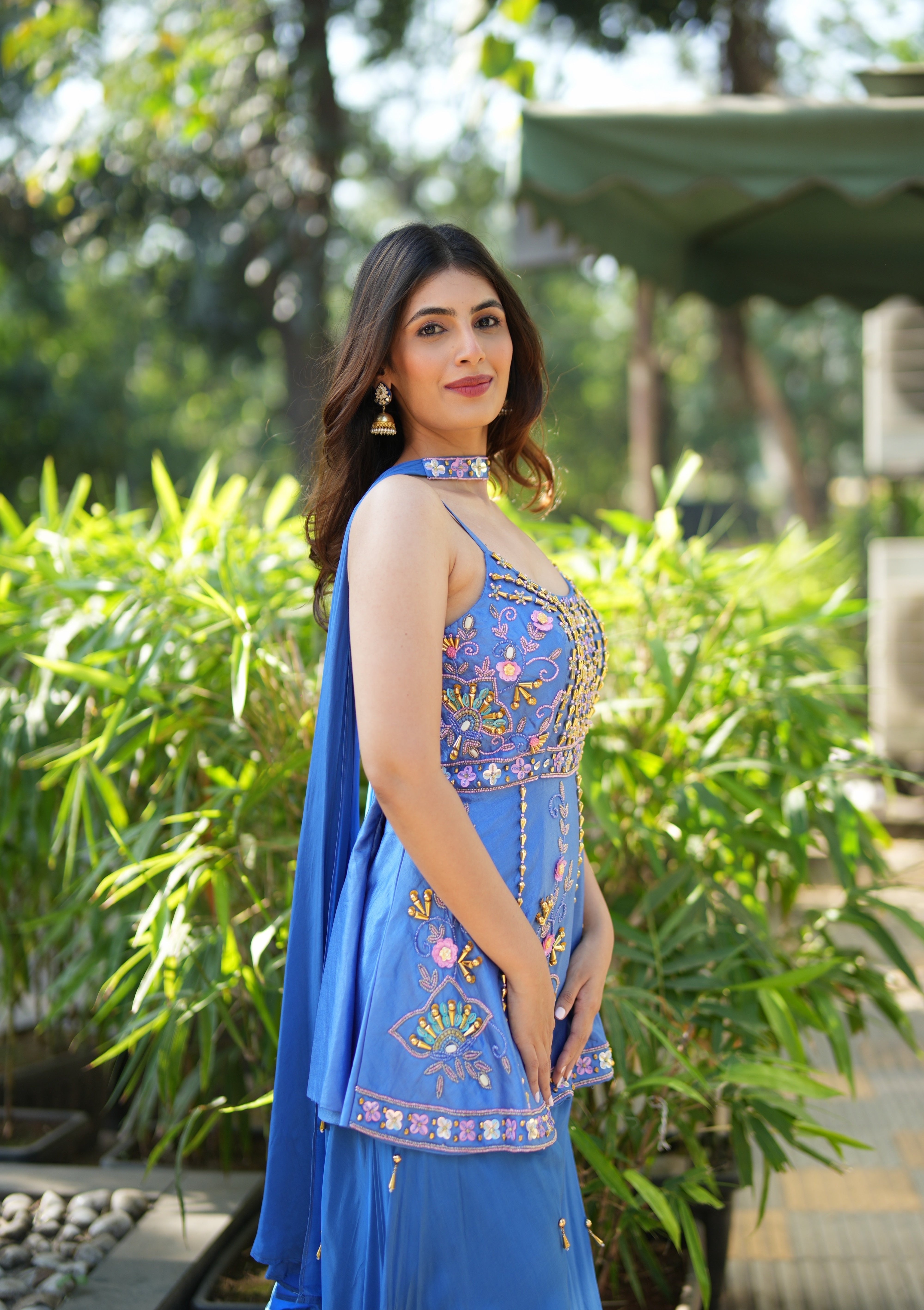 Woman in a blue embroidered outfit standing outdoors with greenery in the background