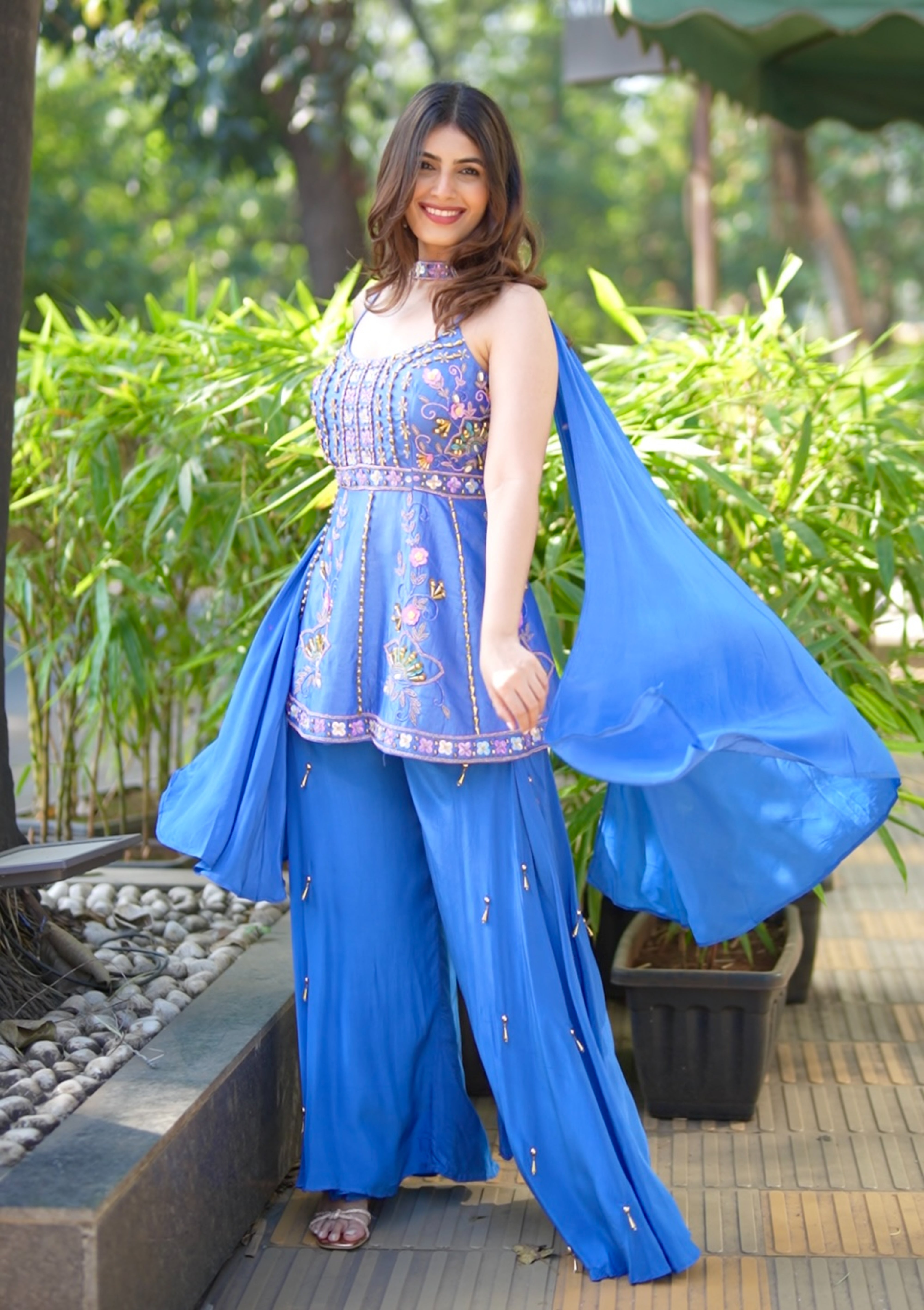 Woman in a blue traditional outfit standing outdoors with greenery in the background