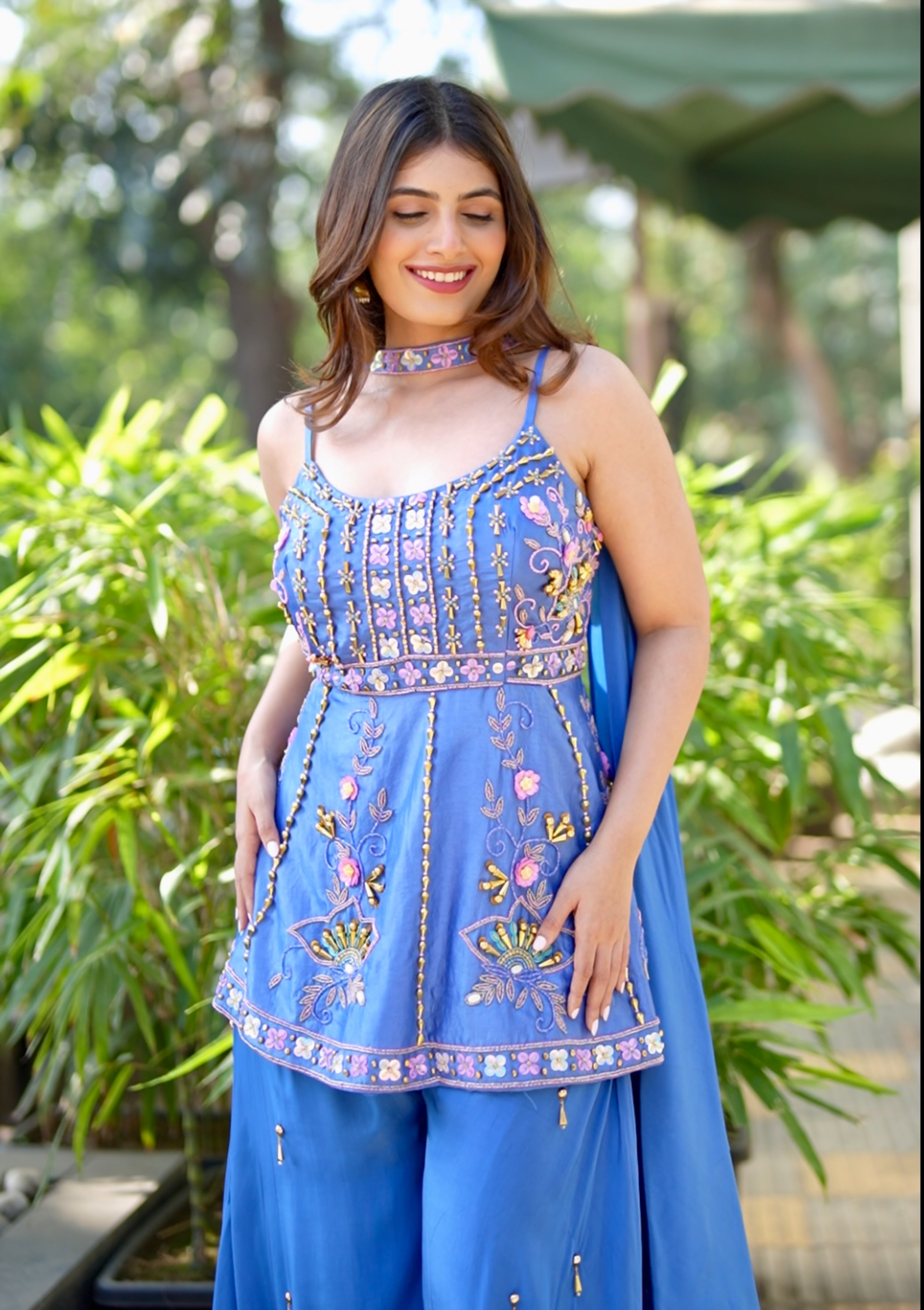 Woman wearing a blue embroidered outfit standing outdoors with greenery in the background
